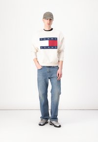 Krémová mikina s námornícky modrým a červeným grafickým nápisom "TOMMY JEANS", skombinovaná so svetlomodrými džínsy a striebornými teniskami. Šedá čiapka nosená nabok.