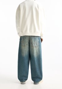 Personne portant un jean en denim bleu surdimensionné et un sweat-shirt blanc, vu de dos sur fond blanc.