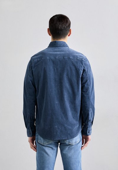 Camisa de manga larga azul marino con cuello abotonado, dobladillo recto y puños de un solo botón, combinada con unos jeans azul claro.