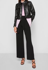 Veste en cuir noir courte avec des fermetures à boutons, associée à une chemise rose pâle et un pantalon noir à taille haute et jambes larges. Talons noirs à lanières.