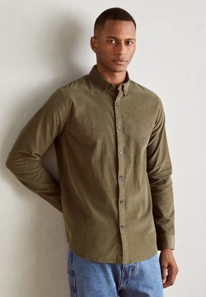 Chemise boutonnée vert olive en tissu texturé avec un col, des manches longues et des boutons ton sur ton discrets, associée à un jean en denim bleu clair.