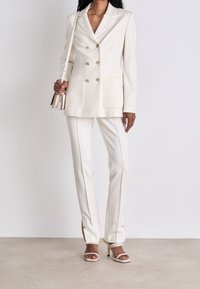 Blazer ivoire à double boutonnage avec quincaillerie dorée, grandes poches, associé à un pantalon ajusté et des talons ouverts avec lanières.