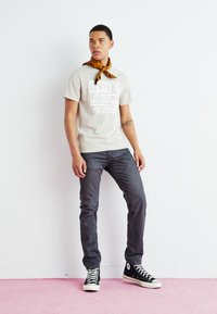 Light gray t-shirt with white graphic text, dark jeans, and black high-top sneakers. Brown patterned scarf tied around the neck.