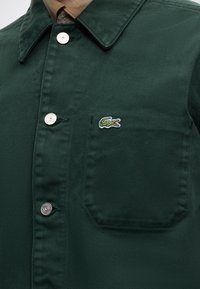 Veste boutonnée vert foncé avec col de chemise marron et petit logo crocodile brodé sur la poche poitrine.