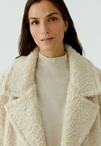 Beige, textured coat with a fluffy exterior; large lapels. Underneath, a light beige ribbed sweater is visible.