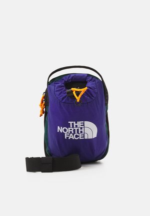 Kompaktowa fioletowo-czarna saszetka North Face z pomarańczowym ściągaczem i czarnym regulowanym paskiem na białym tle.