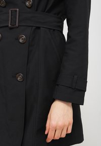 Trench-coat noir double boutonnage avec de grands boutons, une ceinture assortie à boucle rectangulaire, et des manches structurées avec détails à patte.