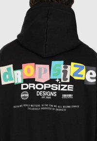 Hoodie noir avec un texte "dropsize" coloré en polices variées, un graphique de globe, un code-barres et la phrase "Rien n'a vraiment d'importance. À la fin, nous devenons tous des histoires."