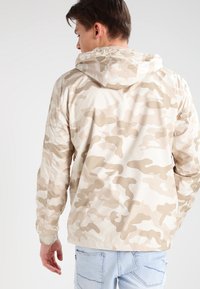 Chaqueta de camuflaje beige con capucha, material ligero, puños y dobladillo elásticos, mostrando una textura suave y un patrón sutil.
