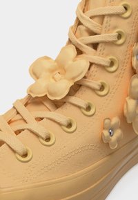 Μπεζ ψηλό αθλητικό παπούτσι με κορδόνια στο ίδιο χρώμα και μεγάλες διακοσμήσεις από υφασμάτινα λουλούδια κοντά στα ρουχιστικά και στην πλευρά.