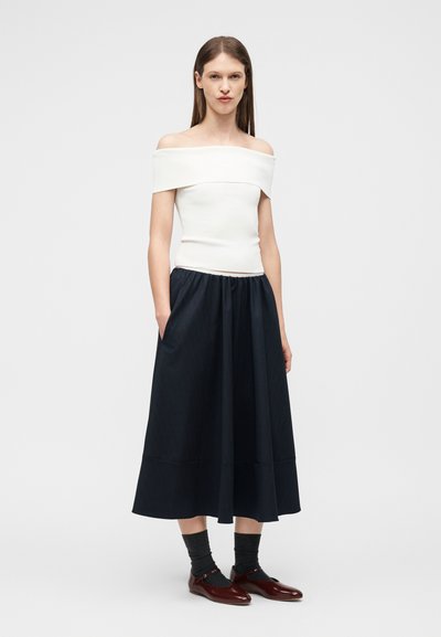 Femme debout portant un haut blanc à épaules dénudées, une jupe midi noire, des chaussettes foncées et des chaussures Mary Jane bordeaux sur un fond clair uni.