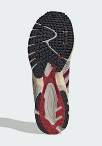 Σόλα αθλητικού παπουτσιού τρεξίματος με μοτίβα πρόσφυσης σε μαύρο, κόκκινο και μπεζ χρώμα, που περιλαμβάνει κυκλικά σχέδια λαβής και την επωνυμία Adidas.