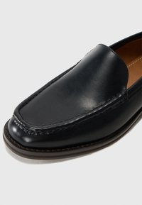 Zapato negro de cuero slip-on con un acabado suave, punta redondeada y detalles de costura visibles. La suela presenta un borde en marrón oscuro contrastante.