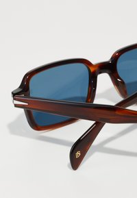 Ochelari de soare tortoiseshell maronii cu lentile albastre. Rame rectangulare cu brațe texturate și finisaj lucios. Logo auriu pe tempă.