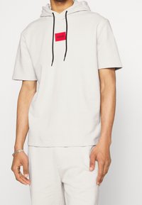 Camiseta con capucha de color gris claro, manga corta, con una etiqueta rectangular roja con el logo "HUGO", cordones negros y un corte holgado.