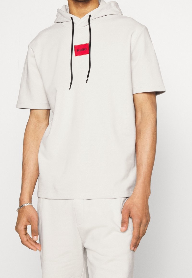 Camiseta con capucha de color gris claro, manga corta, con una etiqueta rectangular roja con el logo "HUGO", cordones negros y un corte holgado.