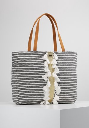 Sac fourre-tout rayé noir et blanc avec des poignées en cuir fauve et une bande verticale dorée ornée de pompons blancs sur une surface blanche.