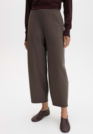 Person trägt cropped weite braune Hosen, dunkelbraunes langärmliges Oberteil und dunkelbraune flache Schuhe und steht vor weißem Hintergrund.