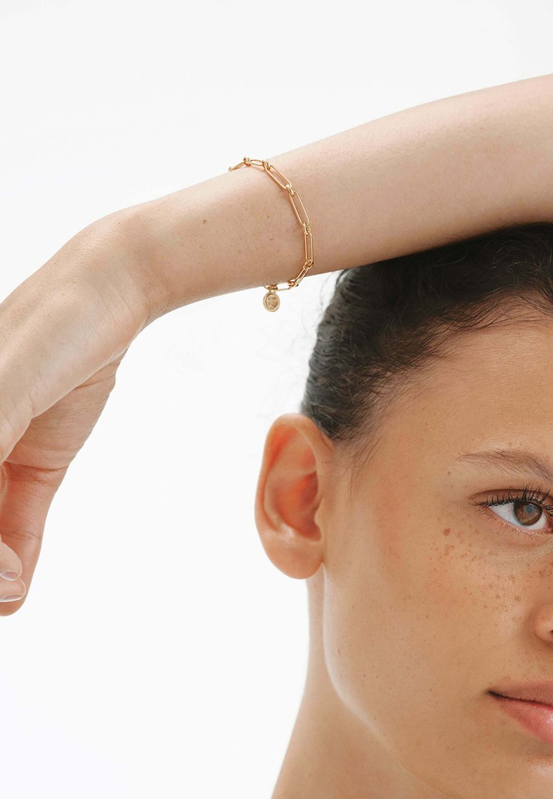 Pulsera de oro con eslabones alargados y un pequeño encanto. Superficie lisa con un acabado pulido, llevada en una mano que descansa sobre un hombro.