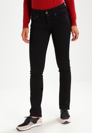 Persoon die zwarte slim-fit jeans draagt, een rood topje met lange mouwen en zwart-roze sneakers, staat met ontspannen handen.