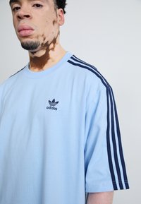Šviesiai mėlynas medvilninis T-marškinėlis su tamsiai mėlynomis trimis juostelėmis ant pečių ir juodu išsiuvinėtu Adidas logotipu ant krūtinės. Trumpomis rankovėmis, laisvo kirpimo.