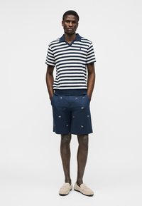 Homme debout, mains dans les poches, portant un polo rayé bleu marine et blanc, un short bleu marine avec de petits imprimés colorés, et des mocassins beiges.