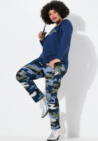 Marineblaues Langarmhemd kombiniert mit blau-grünen Camouflage-Hosen mit Glitzerelementen. Das Model trägt silberne Knöchelboots.