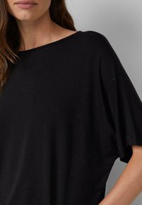 Kobieta w luźnej czarnej bluzce z małymi, subtelnymi błyszczącymi kropkami, odsłaniającej dekolt i część rękawa na szarym tle.