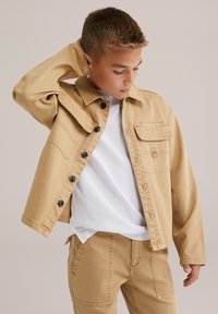 Deček v bež jean jakni in hlačah z belo srajco, ki gleda navzdol z roko za glavo na enotni ozadju.