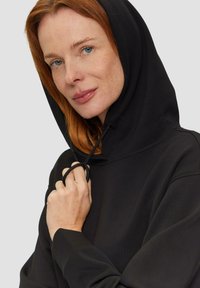 Sudadera con capucha negra y cordón, con una textura suave, un corte relajado y puños acanalados. Primer plano de un modelo sujetando el cordón.