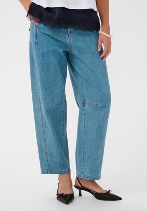 Femme portant un jean bleu clair ample avec des détails cousus aux genoux, des talons slingback noirs pointus, un haut blanc et une bordure en dentelle noire en dessous.