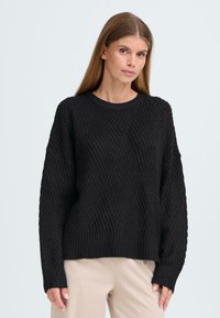 Svart stickad tröja med texturerade diagonala mönster, ribbade ärmmuddar och en klassisk rundad halsringning. Löst sittande med långa ärmar.