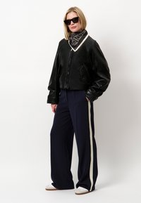 Zwarte leren cropped jack met ritssluiting, gecombineerd met marineblauwe brede broek met een witte zijstreep en een gemusterde zwarte sjaal.