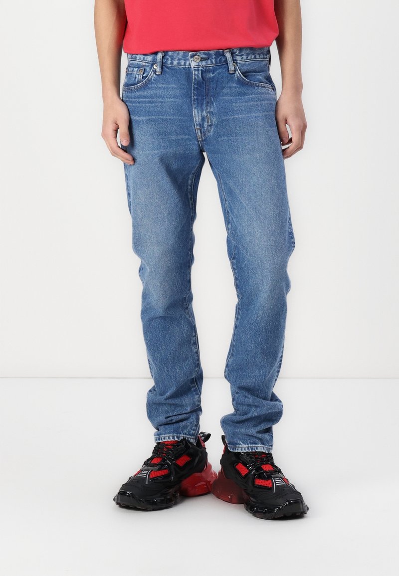 Jean en denim bleu clair avec une coupe droite et une texture délavée, associé à des chaussures de sport noires et rouges à semelle épaisse.