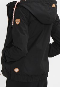 Svart jacka med huva, utrustad med elastisk midja, texturerad tyg och logopatchar på axlarna. Inkluderar flätade dragskor.