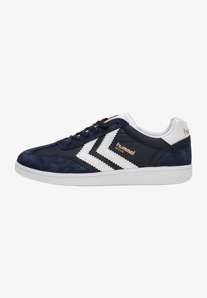 Zapatilla Hummel azul marino y blanca con cierre de cordones, franjas blancas en forma de cheurón, detalles de ante y suela de goma blanca texturizada.