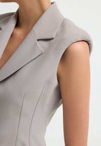 Hellegraue ärmellose Blazerjacke mit strukturierten Schultern, scharfen Reverskragen und vertikalen Nahtdetails. Glatte Stofftextur sichtbar.