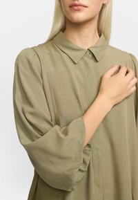 Camicia verde oliva con colletto, chiusura a bottoni e maniche a sbuffo. Tessuto liscio con una sottile trama.