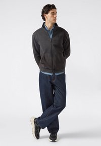Hombre de pie con los brazos detrás de la espalda, vistiendo un forro polar negro con cremallera, una camisa azul de botones, jeans oscuros y zapatillas negras y blancas, sobre un fondo blanco.
