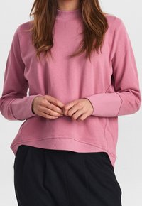 Rosa Sweatshirt mit hohem Kragen, gerippten Bündchen und lockerer Passform. Die Textur wirkt weich und leicht elastisch. Getragen mit schwarzen Hosen.