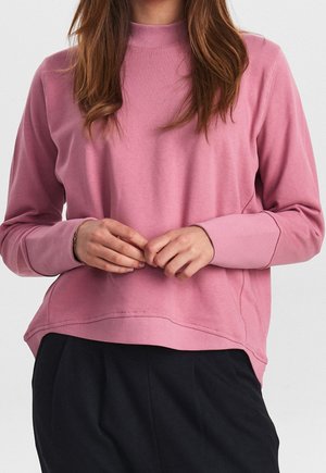 Felpa rosa con un collo alto, polsini a coste e una vestibilità rilassata. La texture appare morbida e leggermente elastica. Indossata con pantaloni neri.
