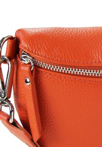 Sac en cuir orange avec une finition texturée. Comprend une fermeture éclair argentée et une bandoulière détachable ornée de détails métalliques.