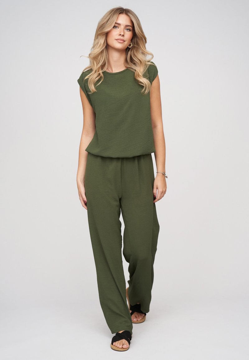 Femme aux longs cheveux blonds porte un haut sans manches vert olive et un pantalon large assorti avec des sandales noires sur fond clair.
