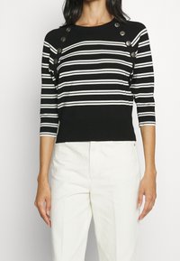 Kobieta ubrana w czarną i białą poziomo paskowaną bluzkę z srebrnymi guzikami na ramionach oraz białe spodnie z wysokim stanem.