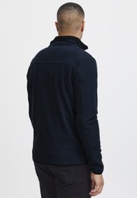 Marineblå fleece-pullover med høy krave, med en teksturert overflate, lange ermer og kontrasterende svarte detaljer ved kragen.