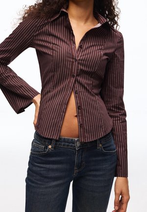 Kobieta ubrana w dopasowaną, brązową koszulę w cienkie paski z długim rękawem, luźno rozpiętą na ciemnoniebieskich dżinsach na jednolitym tle.