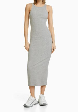 Femme portant une robe midi grise, moulante et sans manches, en maille côtelée, associée à des baskets blanches épaisses, debout devant un fond blanc uni.