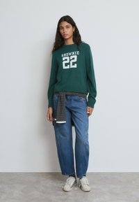 Suéter verde con el texto blanco "BROWNIE 22", combinado con jeans de corte relajado en azul. Material de punto texturizado, diseño casual y detalle a rayas en la cintura.