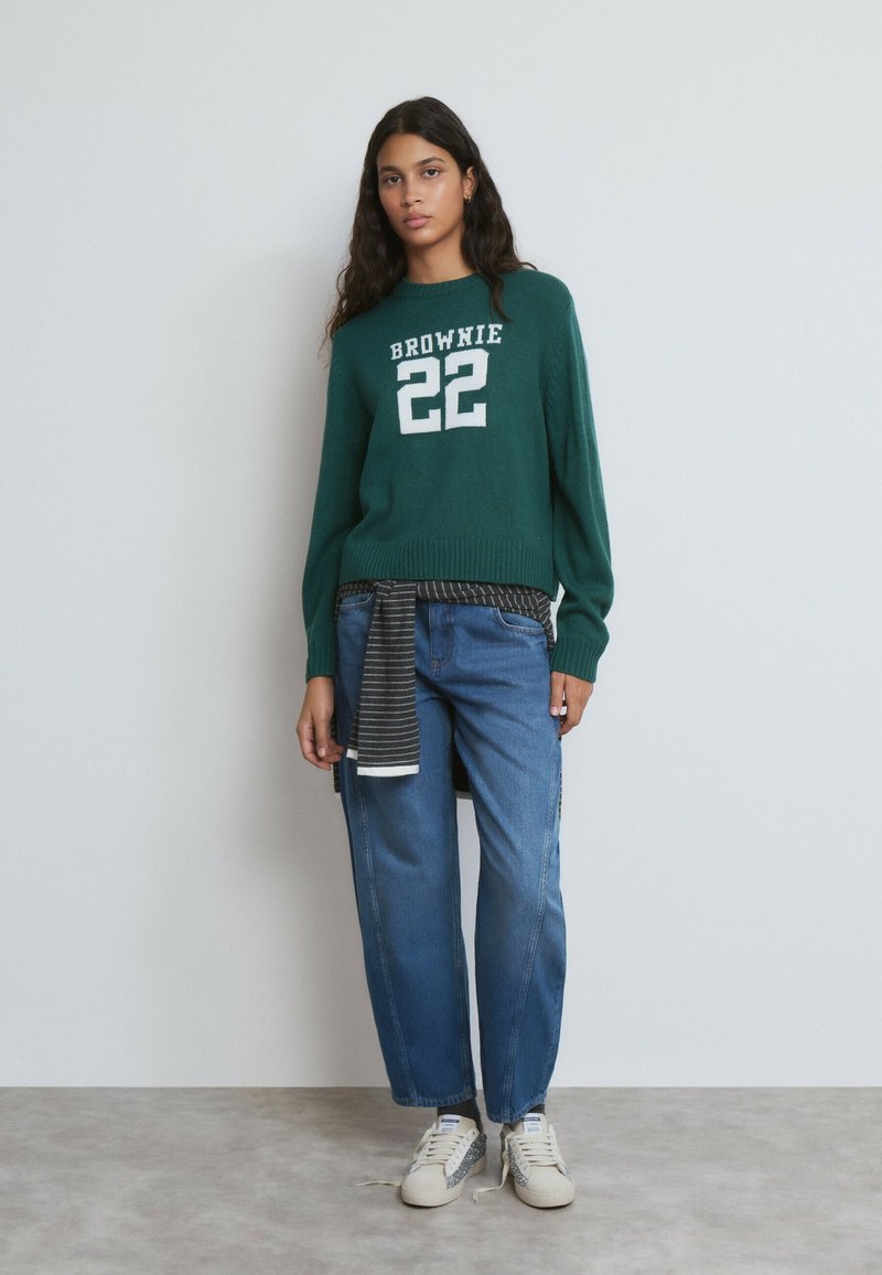 Suéter verde con el texto blanco "BROWNIE 22", combinado con jeans de corte relajado en azul. Material de punto texturizado, diseño casual y detalle a rayas en la cintura.
