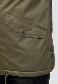 Zielona kurtka z tkaniny z boczną kieszenią z zatrzaskiem, przeszyty szew i sznurek w dole. Teksturowana powierzchnia.